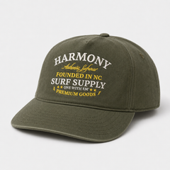 An olive green baseball cap with embroidered text on the front reading 'HARMONY Authentic Lifestyle FOUNDED IN NC SURF SUPPLY ONE WITH EM PREMIUM GOODS' in white and yellow, shown on a light gray background.