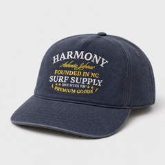 A navy blue baseball cap with white and yellow embroidered text on the front reading 'HARMONY Authentic Lifestyle FOUNDED IN NC SURF SUPPLY ONE WITH EM PREMIUM GOODS'.