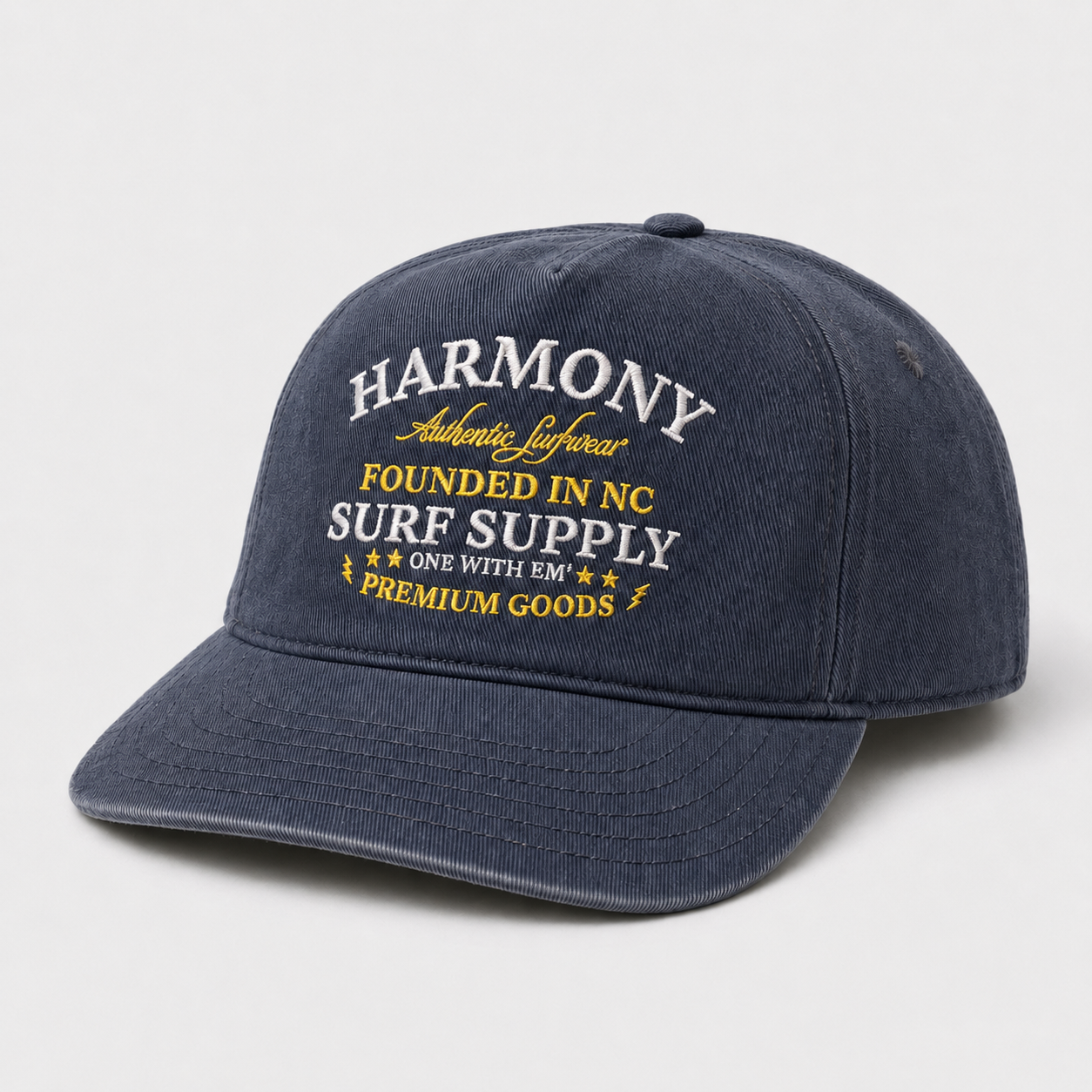 A navy blue baseball cap with white and yellow embroidered text on the front reading 'HARMONY Authentic Lifestyle FOUNDED IN NC SURF SUPPLY ONE WITH EM PREMIUM GOODS'.