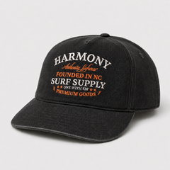 A black washed baseball cap with embroidered text on the front reading 'HARMONY Authentic Lifestyle FOUNDED IN NC SURF SUPPLY ONE WITH EM PREMIUM GOODS' in white and orange, shown on a light gray background.