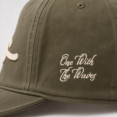 Close-up of an olive green baseball cap with cream-colored embroidered text reading 'One With The Waves' on the front panel.