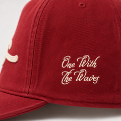 A close-up of a red baseball cap featuring white embroidered text that reads 'One With The Waves' on the side panel, with a partial white embroidered logo visible on the front.