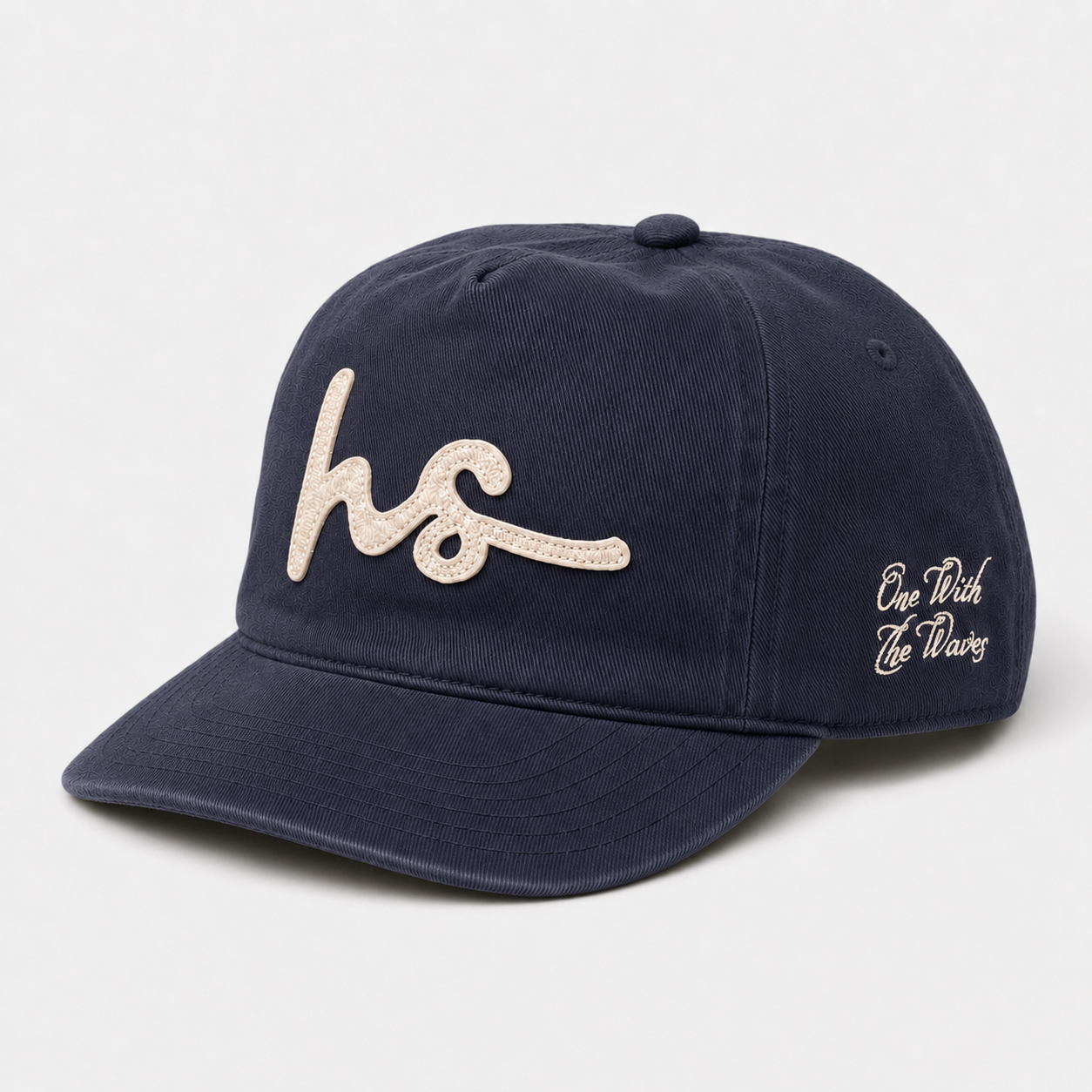 A navy blue cotton baseball cap featuring a large, textured beige embroidered 'hs' logo on the front and 'One With The Waves' in beige script on the side, set against a light gray background.