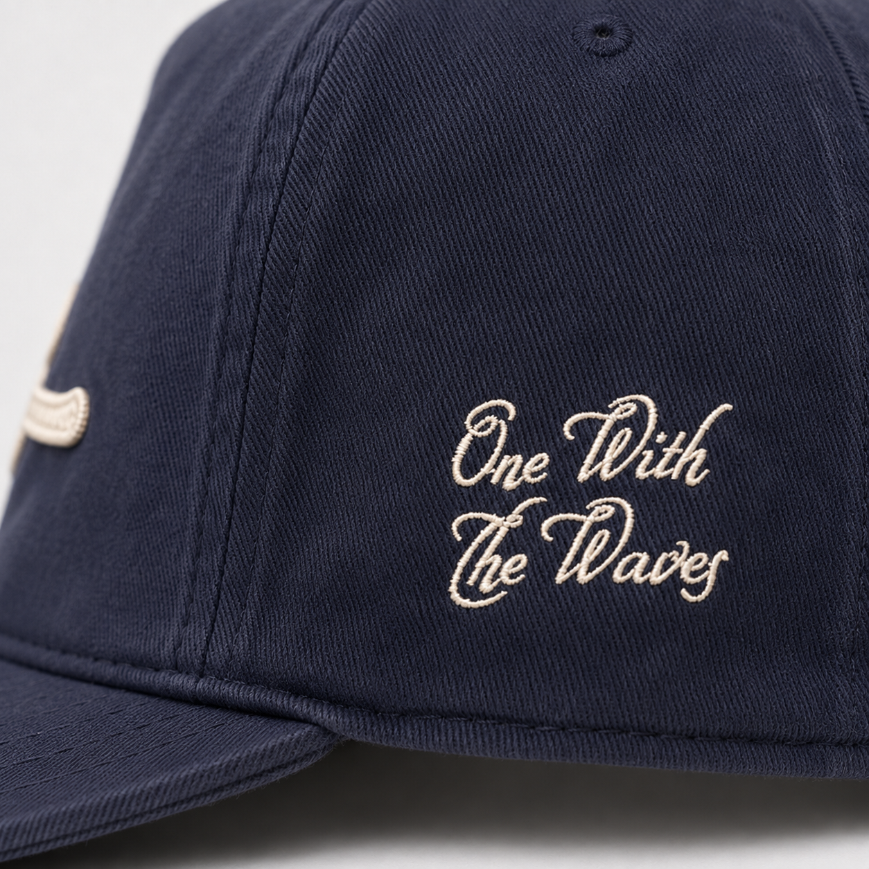 Close-up of a navy blue baseball cap with white embroidered text reading 'One With The Waves' on the front panel.