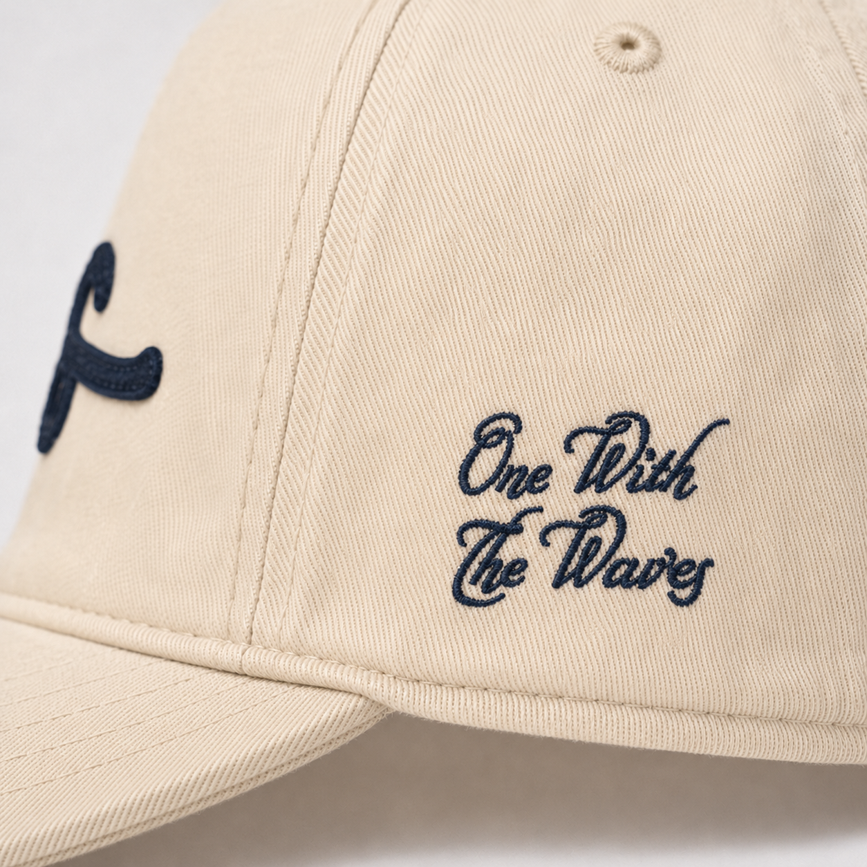 Close-up of a beige baseball cap with navy blue embroidered text reading 'One With The Waves' on the side panel and a matching navy script logo on the brim.